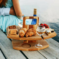 Wooden table on a lakeside jetty, loaded with croissants, nuts, fruit and wine, with a seated woman behind 2