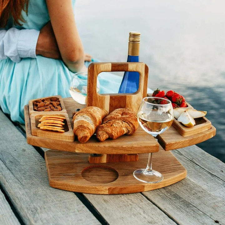Wooden table holding croissants, nuts, crackers, fruit and wine, set on a jetty beside a woman in turquoise 4