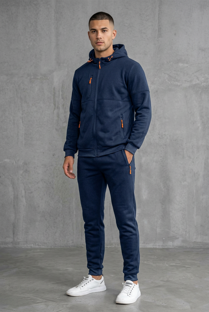 Navy hooded zip tracksuit with orange zip pulls, worn by a model, front view against concrete wall 2