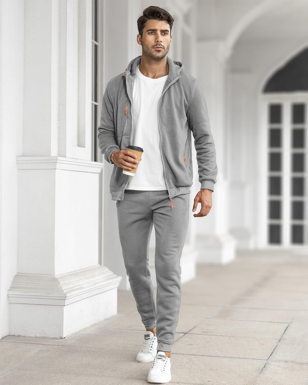 Model walking through a white colonnade in the grey zip-up tracksuit with a white t-shirt, holding a coffee 30