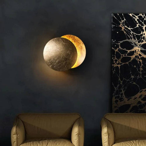 Gold-toned disc lamp glowing on a dark wall above two olive armchairs, beside marbled artwork 2