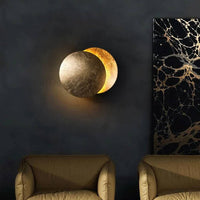 Lamp with a textured gold front disc on a dark wall above two armchairs, beside marbled artwork 3