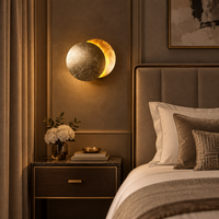 Gold-leaf moon wall light glowing above a marble-topped bedside table in a warm, neutral bedroom 1