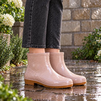 Glossy pink ankle rain boots with tan soles worn with dark jeans, speckled with raindrops on a wet paved path. 1