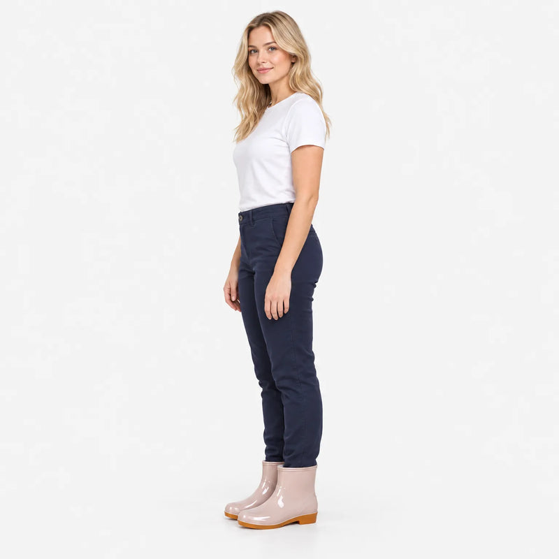 Woman in white t-shirt and navy trousers standing in profile wearing pink glossy ankle boots against a plain white background. 4
