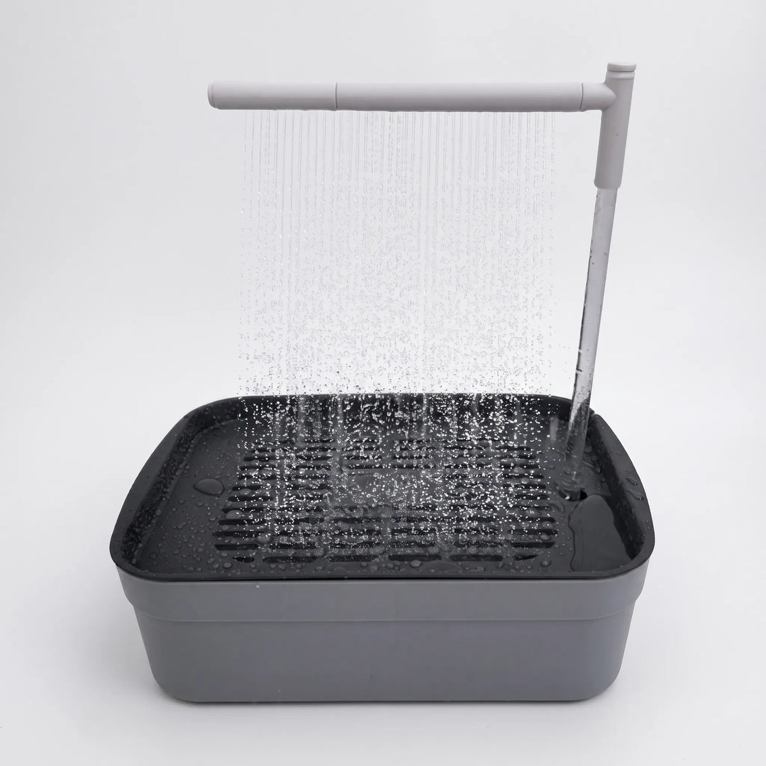 Grey bird bath with a dark slatted tray, water pouring from an overhead arm as a fine shower on a plain white background. 1