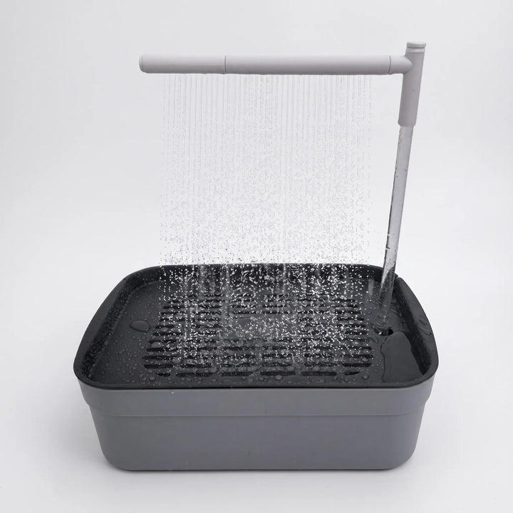 Grey bird bath with a dark slatted tray, water pouring from an overhead arm as a fine shower on a plain white background. 1
