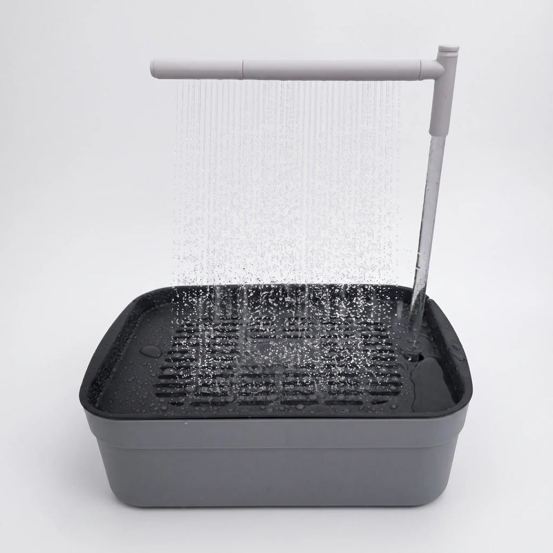 Grey bird bath with a dark slatted tray, water pouring from an overhead arm as a fine shower on a plain white background. 1
