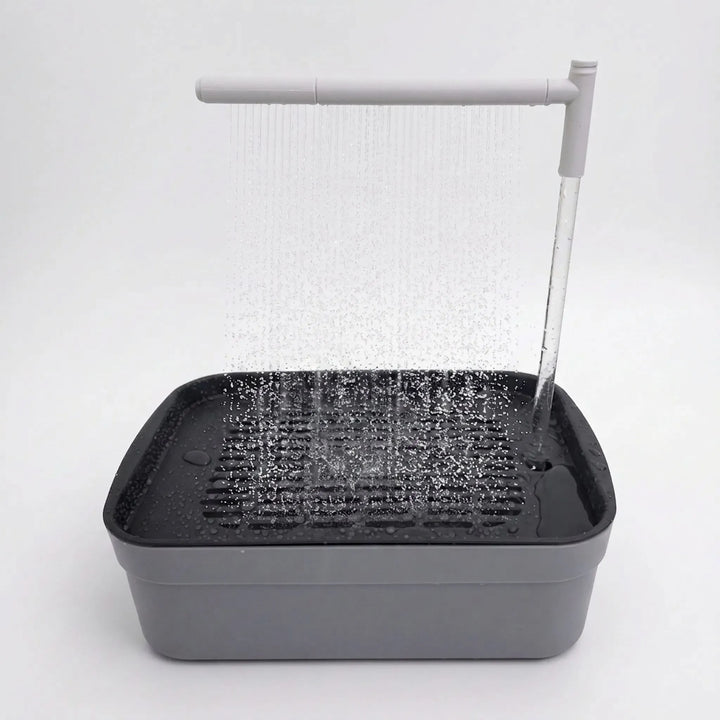 Grey bird bath on a plain white background with water falling from an overhead spray arm onto the slatted tray. 1