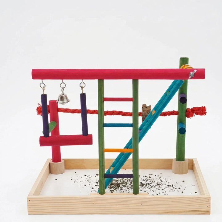 Front view of the colourful wooden bird playground on a plain white background, showing swing, bell, ladder, rope perch and seed tray. 1