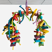 Arched toy hung from a cage roof, with coloured wooden blocks, beads, braided cotton rope strands and small brass bells on a white background. 1