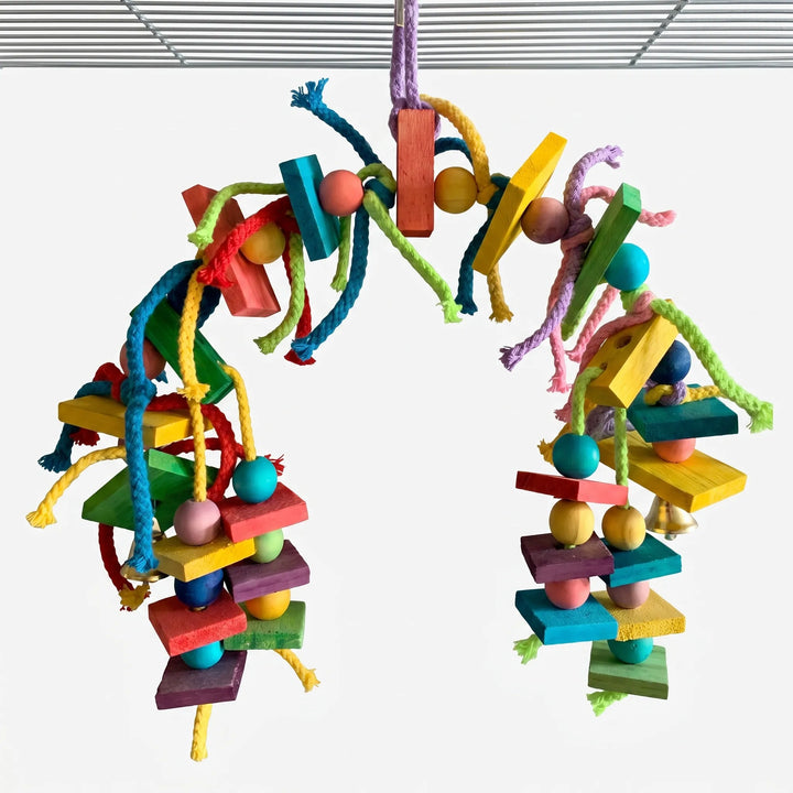 Arched toy hung from a cage roof, with coloured wooden blocks, beads, braided cotton rope strands and small brass bells on a white background. 1