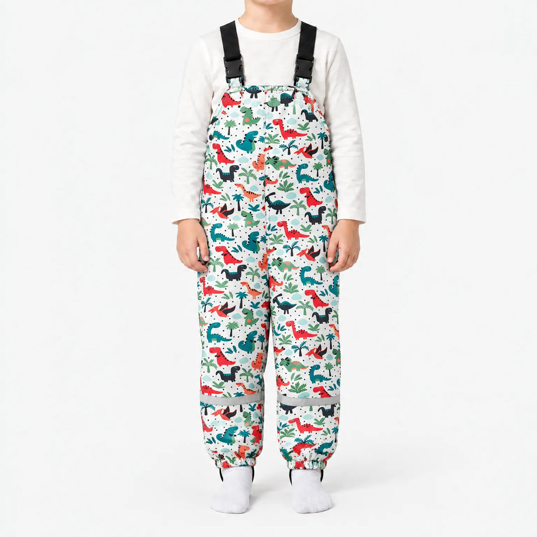 Front view of white dungarees with a pattern of red, teal and black dinosaurs among palm trees, worn with a white top. 12