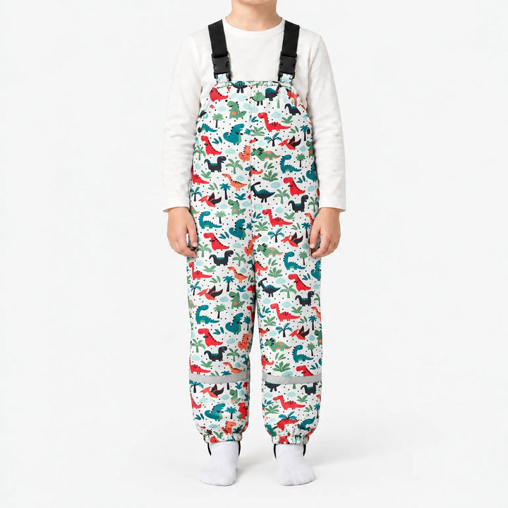 Front view of white dungarees with a pattern of red, teal and black dinosaurs among palm trees, worn with a white top. 12