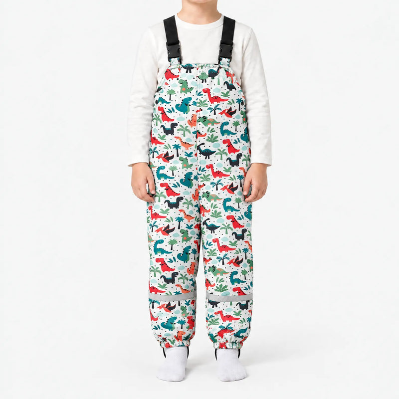 Front view of white dungarees with a pattern of red, teal and black dinosaurs among palm trees, worn with a white top. 12