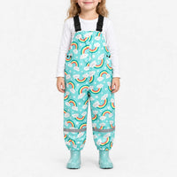 Smiling girl wearing turquoise dungarees printed with smiling clouds, rainbows and stars, with matching turquoise wellies and a white top. 2