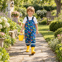 Smiling boy in blue dinosaur-print dungarees and yellow wellies walking along a sunny garden path carrying a yellow bucket. 3