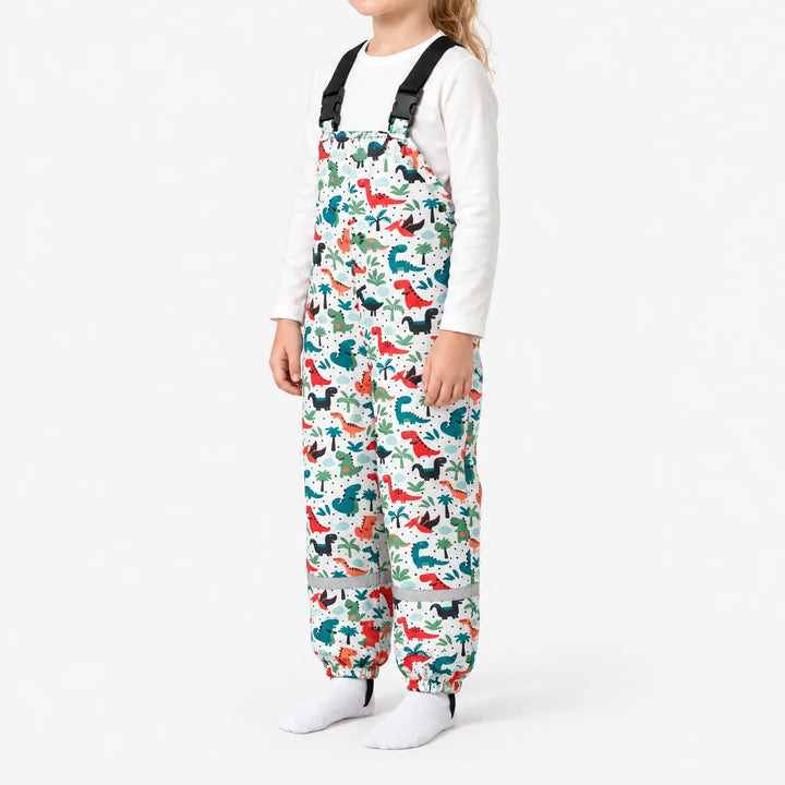Side view of a child wearing white dungarees with a colourful dinosaur and palm tree print, reflective band at the ankles. 5