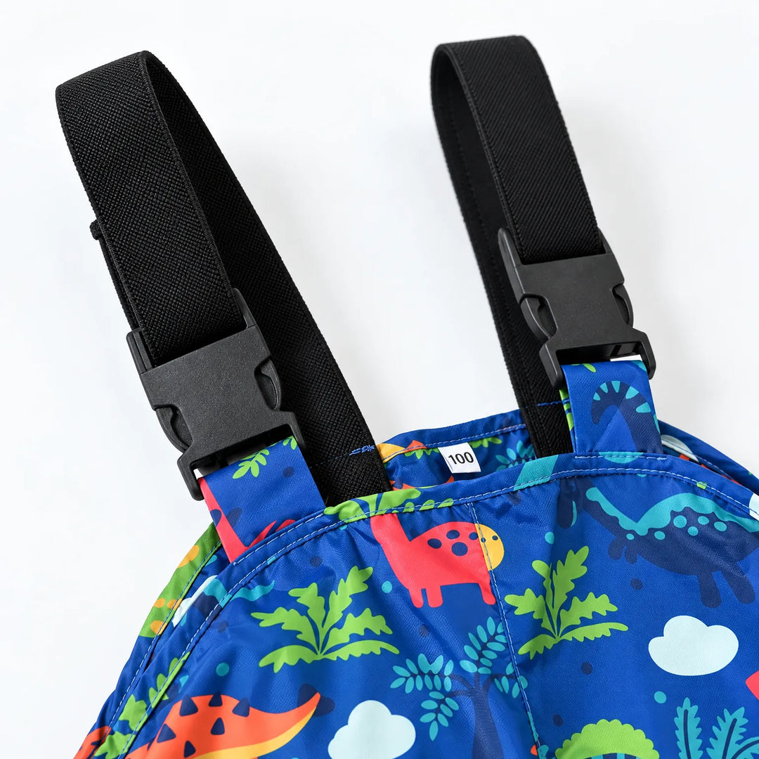 Close-up of the black adjustable shoulder straps with plastic buckles on blue dinosaur-print dungarees, with a small size label inside. 6