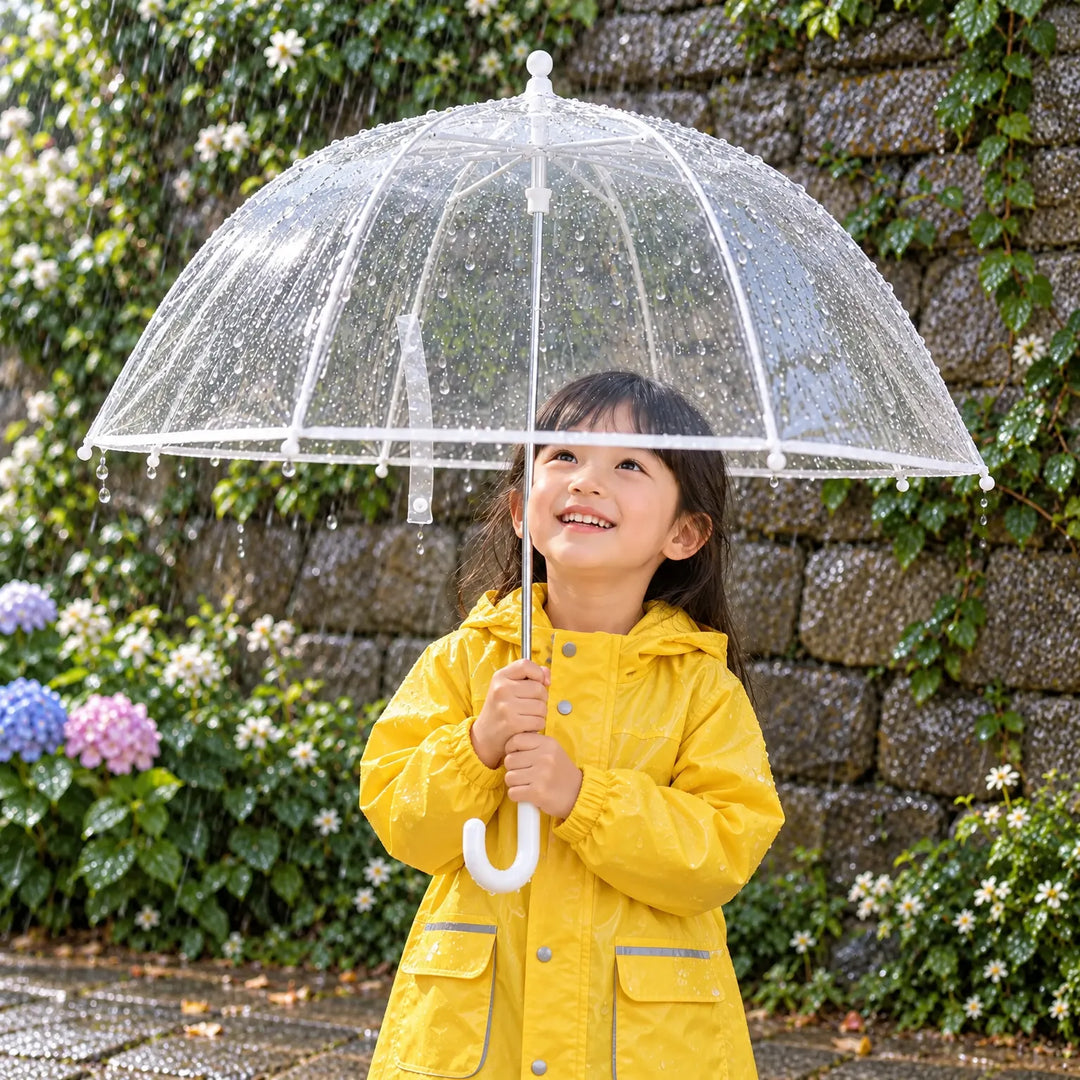 Smiling girl in a yellow raincoat holds the clear dome umbrella with rain droplets, in front of a stone wall and hydrangeas. 1