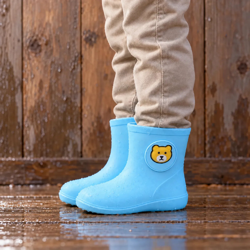 Child wearing light blue rain boots with a round bear badge, standing on a wet wooden floor with water droplets. 1