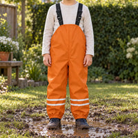 Child in orange waterproof dungarees with reflective leg stripes standing in a muddy puddle on a garden lawn in sunshine. 1