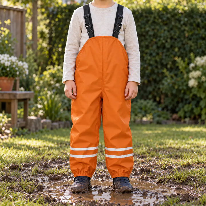 Child in orange waterproof dungarees with reflective leg stripes standing in a muddy puddle on a garden lawn in sunshine. 1