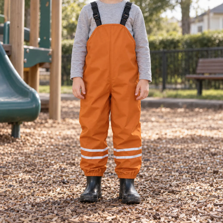 Child in burnt orange waterproof dungarees and black wellies standing on wood chips at a playground, seen from the front. 3