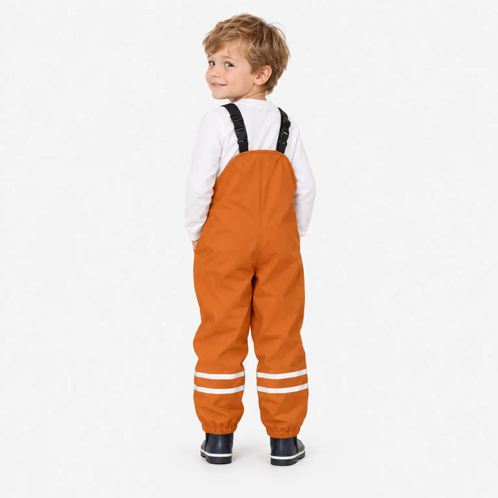 Smiling boy seen from behind in orange waterproof dungarees, looking over his shoulder, showing the plain back and elastic ankle cuffs. 4