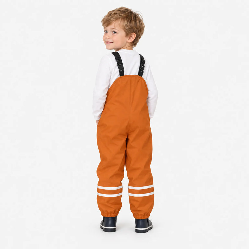 Smiling boy seen from behind in orange waterproof dungarees, looking over his shoulder, showing the plain back and elastic ankle cuffs. 4