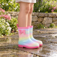 Child standing in rainbow-gradient wellies with a pink bow on a wet stone path beside a flower bed. 1