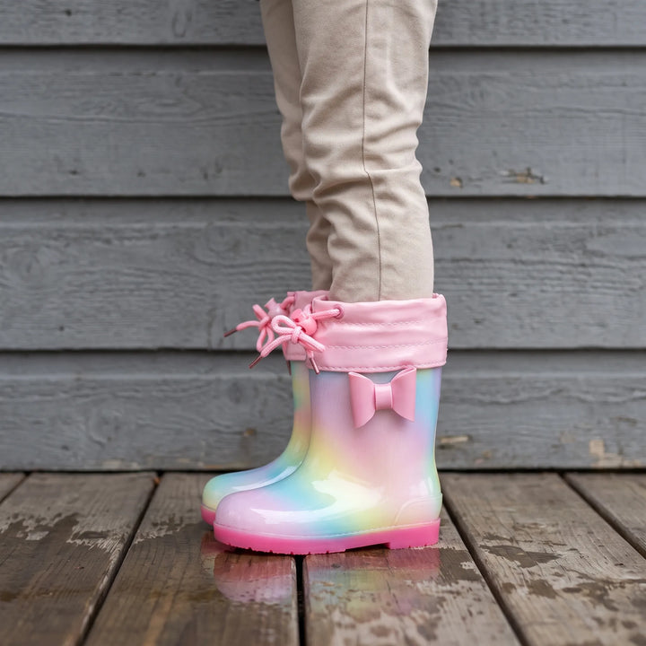 Child in beige trousers wearing rainbow wellies with a pink drawstring cuff and bow, standing on a wooden deck. 3