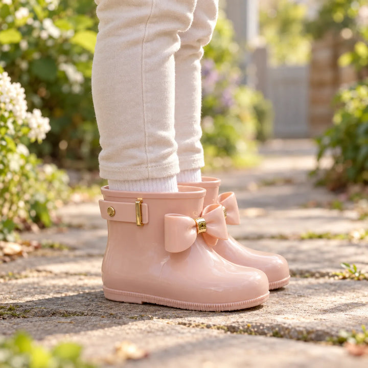 Side view of a child wearing glossy blush pink wellies with a bow and gold buckle strap, standing on a garden path. 1