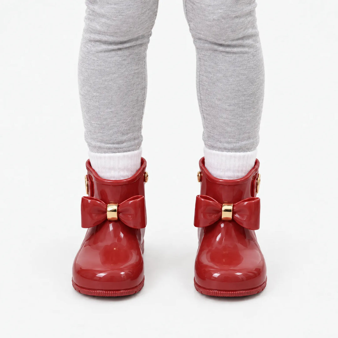Front view of a child in grey leggings and white socks wearing glossy red wellies with matching bows and gold bands. 10