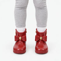 Front view of a child in grey leggings and white socks wearing glossy red wellies with matching bows and gold bands. 10