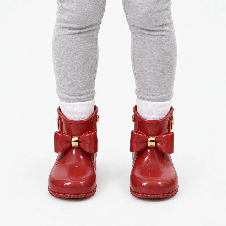 Front view of a child in grey leggings and white socks wearing glossy red wellies with matching bows and gold bands. 10