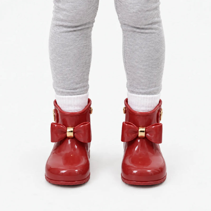 Front view of a child in grey leggings and white socks wearing glossy red wellies with matching bows and gold bands. 10