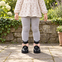 Child in grey leggings and a floral top stands in a garden wearing black wellies with pink bows and gold details. 3