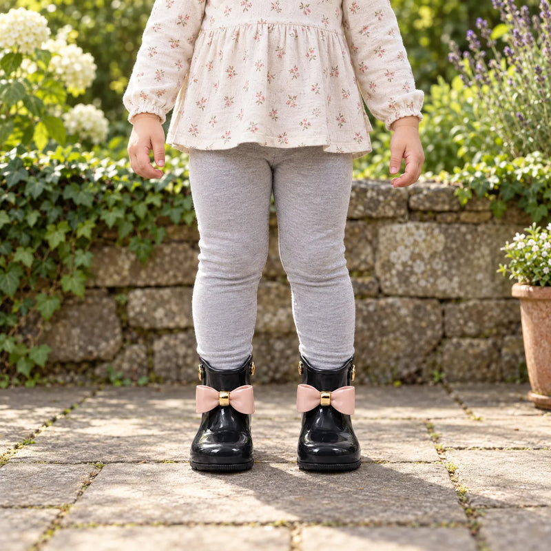 Child in grey leggings and a floral top stands in a garden wearing black wellies with pink bows and gold details. 3