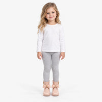 Smiling girl with long wavy hair in a white top and grey leggings, wearing blush pink wellies with bows, full-length studio shot. 4