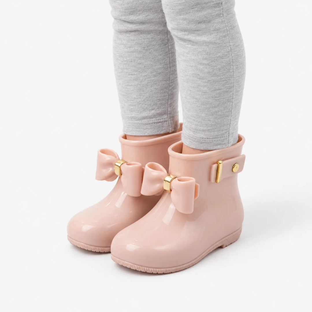 Angled view of a child's legs in grey leggings wearing blush pink wellies with bows, showing the side strap and gold buckle. 5