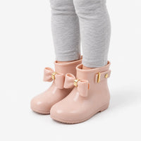 Angled view of a child's legs in grey leggings wearing blush pink wellies with bows, showing the side strap and gold buckle. 5