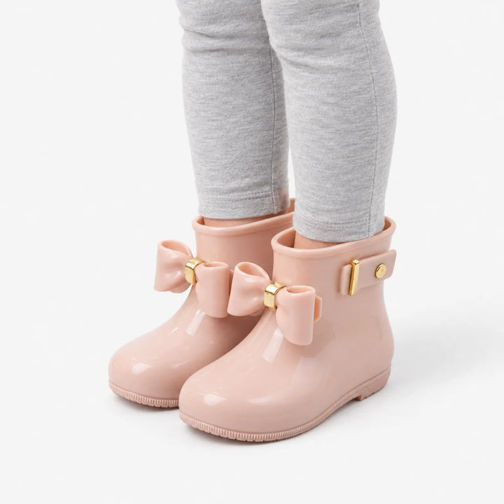Angled view of a child's legs in grey leggings wearing blush pink wellies with bows, showing the side strap and gold buckle. 5