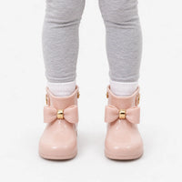 Front view of a child in grey leggings and white socks wearing blush pink wellies with gold-banded bows against white. 7