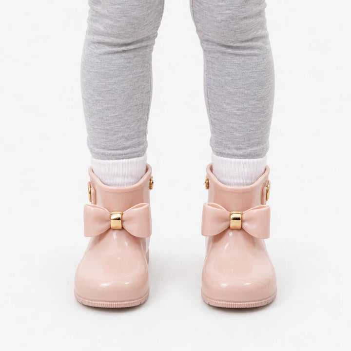 Front view of a child in grey leggings and white socks wearing blush pink wellies with gold-banded bows against white. 7