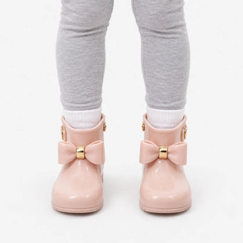 Front view of a child in grey leggings and white socks wearing blush pink wellies with gold-banded bows against white. 7