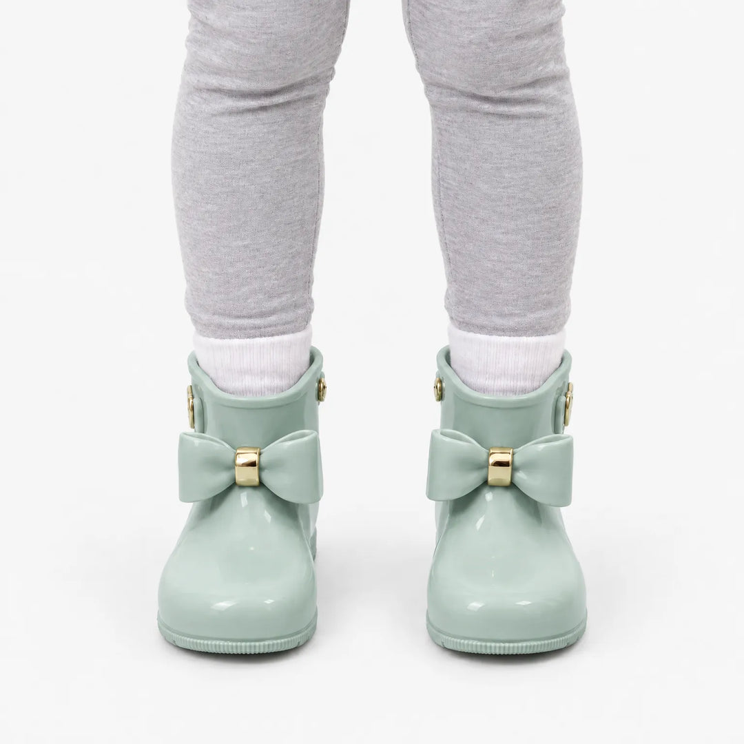 Front view of a child in grey leggings and white socks wearing sage green wellies with matching bows and gold bands. 8