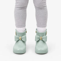 Front view of a child in grey leggings and white socks wearing sage green wellies with matching bows and gold bands. 8