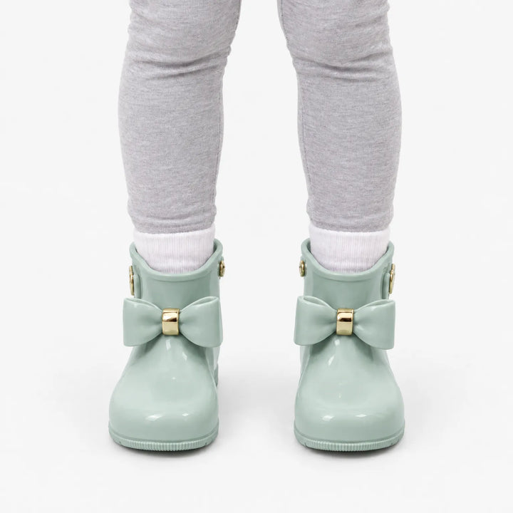 Front view of a child in grey leggings and white socks wearing sage green wellies with matching bows and gold bands. 8