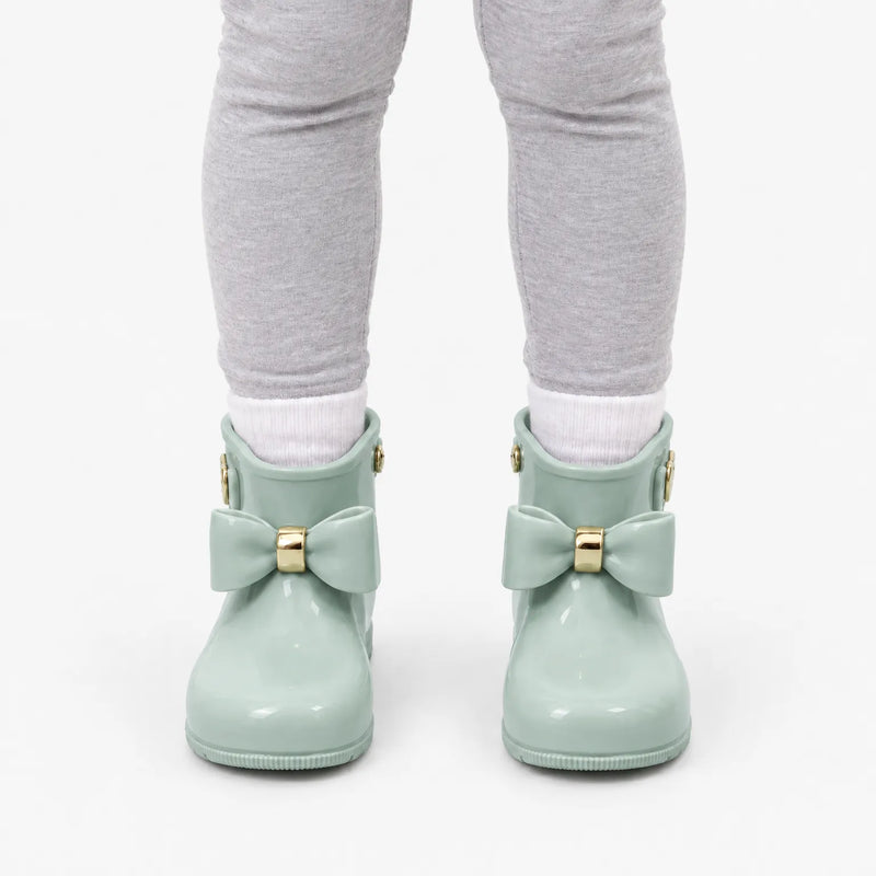 Front view of a child in grey leggings and white socks wearing sage green wellies with matching bows and gold bands. 8